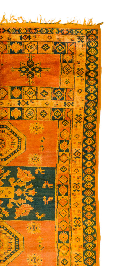 A close-up of a patterned rug showing orange, mustard yellow, and dark green geometric and floral motifs with fringe along the top edge.