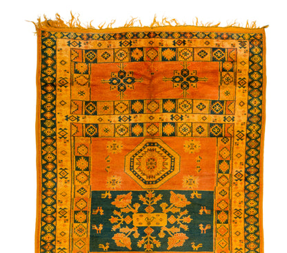 An antique-style geometric area rug with a warm orange and mustard yellow field, dark green borders, and a central teal section with ornate floral motifs and fringed edges.
