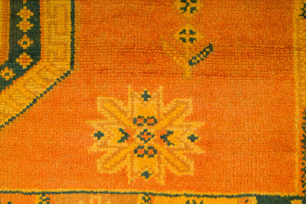 Close-up of an orange patterned rug with a central floral medallion and dark green and yellow geometric borders.