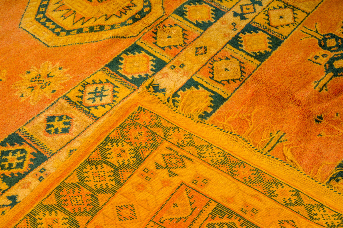 A close-up of a patterned rug featuring a warm orange and yellow base with dark green geometric borders and motifs.