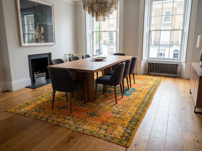 A dining room scene with a rectangular wooden dining table and six dark upholstered chairs placed on a large patterned area rug. The room has light wood floors, tall windows, a fireplace, and a chandelier overhead.
