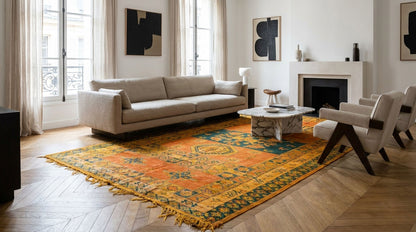 A large vintage-style oriental area rug with orange, yellow, and blue patterns and fringed edges, styled in a bright, modern living room with a beige sofa and wooden floors.