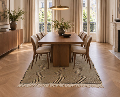 A dining room scene with a rectangular wooden table surrounded by six upholstered chairs on a patterned beige Moroccan Zanafi rug with fringe, set in a bright room with tall windows, curtains, and wood flooring.