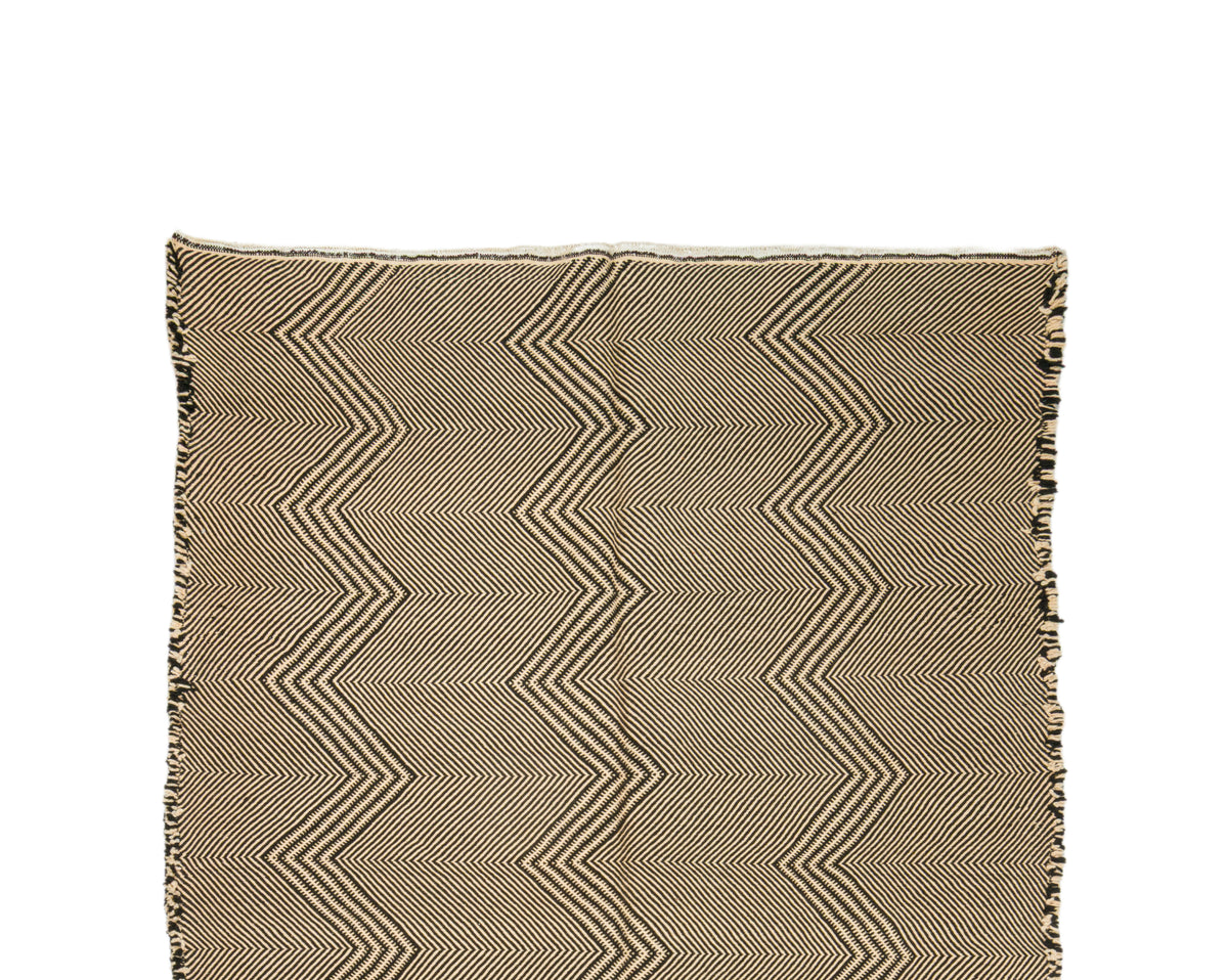 Detail of a Zanafi rug with fringe edges, featuring a beige and brown zigzag pattern on a white background.