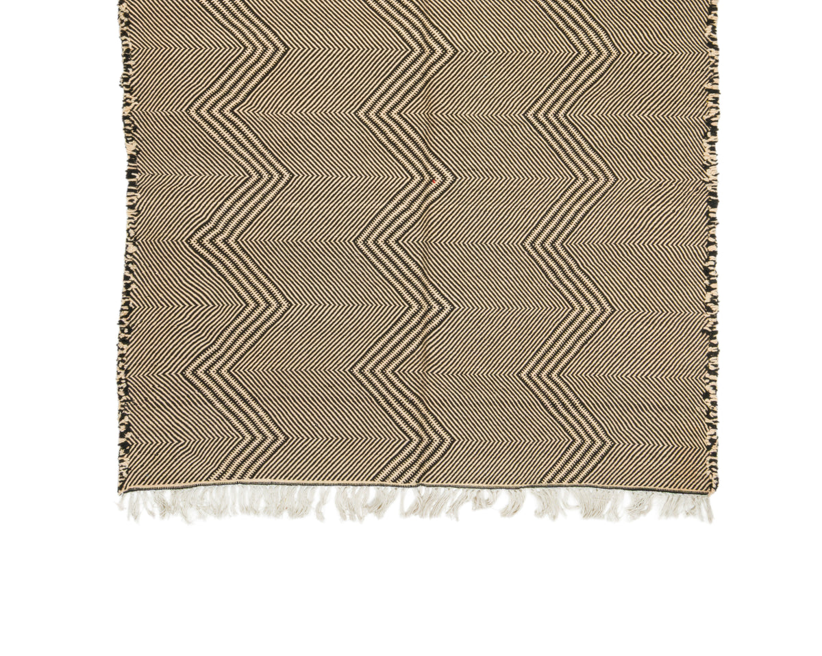 A woven Moroccan rug with a beige and brown chevron pattern and white fringe tassels, shown flat against a white background.