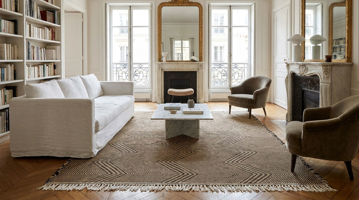 A large wool Zanafi rug from Morocco with a geometric chevron pattern and fringe, styled in a bright, classic Parisian living room with a white sofa, armchairs, bookshelves, and fireplace.