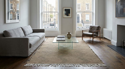 A Moroccan Zanafi rug in a modern living room featuring the large beige and brown geometric patterned area rug with fringe, styled with a gray sofa, a glass-top coffee table, and a wooden armchair.