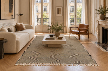 A large Moroccan Zanafi rug with a beige and brown geometric chevron pattern and fringe, styled in a bright, modern living room with a cream sofa, wooden coffee table, and herringbone wood floors.