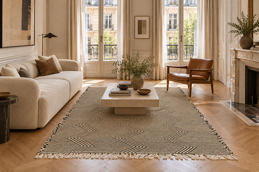 A large Moroccan Zanafi rug with a beige and brown geometric chevron pattern and fringe, styled in a bright, modern living room with a cream sofa, wooden coffee table, and herringbone wood floors.