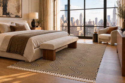 A styled bedroom interior featuring a large woven area rug with a geometric chevron pattern and fringe, placed under a bed with a matching upholstered bench. The room has warm wood floors, beige curtains, a city skyline view through large windows, and a cream armchair.