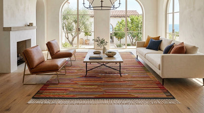 A styled living room featuring a prominent multicolor striped Moroccan area rug with fringe, styled with a cream sofa, two brown leather armchairs, and a small wooden coffee table.