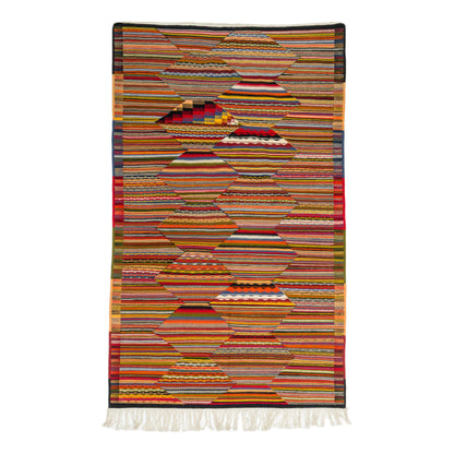 A rectangular woven Moroccan  rug with a geometric pattern made of horizontal striped textile sections in many colors, finished with white fringe along the bottom, shown on a plain white background.