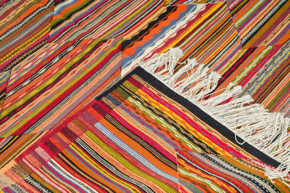 A close-up of a colorful woven textile with dense diagonal stripes in orange, red, pink, yellow, green, blue, black, and white, finished with white fringe on one end.