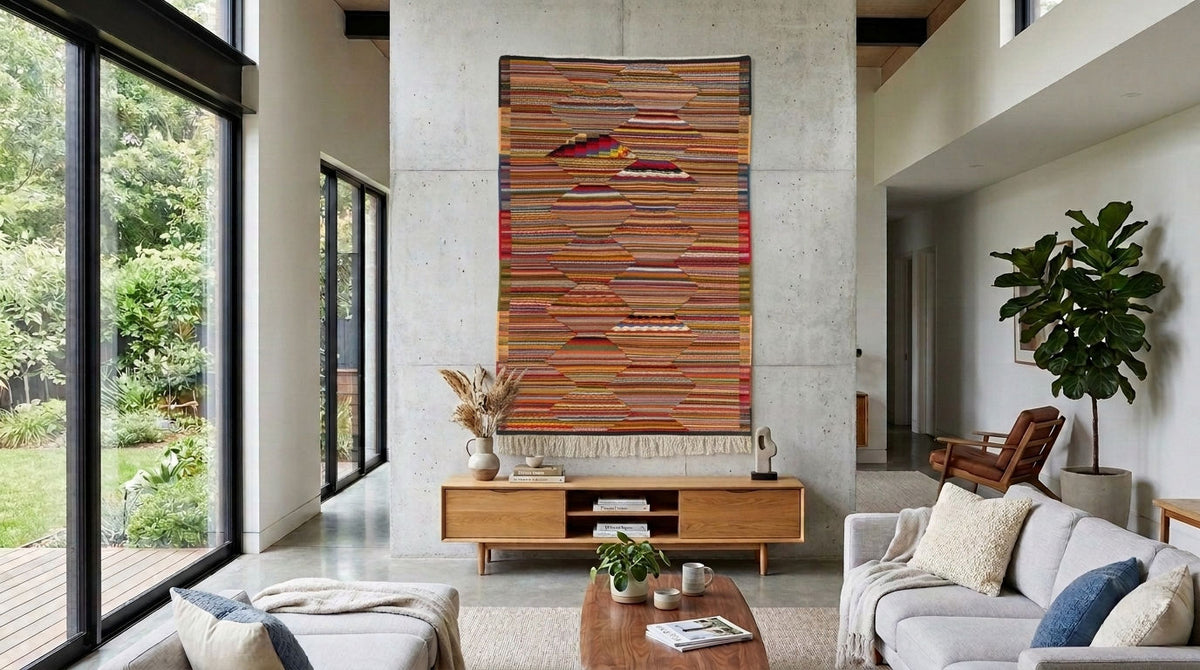 A large rectangular woven wall hanging with horizontal multicolor stripes and fringe at the bottom, displayed in a modern living room above a wooden console table.