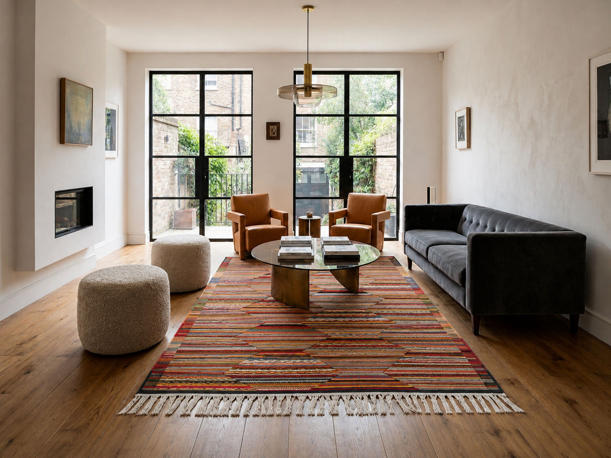 A modern living room featuring a prominent striped Moroccan area rug with cream fringe, styled with a dark gray sofa, two tan armchairs, and two round poufs on a hardwood floor.