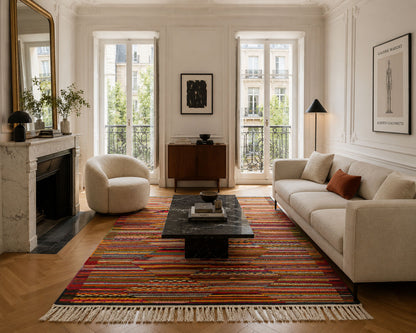 A styled living room interior featuring a large multicolor striped area rug with cream fringe, a cream sofa, a matching armchair, a black coffee table, and a white marble fireplace.