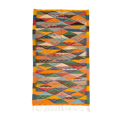 A rectangular handwoven cubist moroccan  rug with a vibrant geometric diamond pattern in orange, yellow, green, blue, red, and brown, finished with white fringe at the bottom, shown flat on a white background.