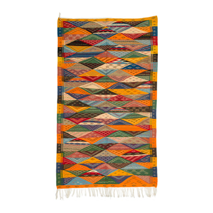 A colorful geometric woven moroccan rug with a diamond pattern, featuring orange borders and white fringe at the bottom, shown against a plain white background.