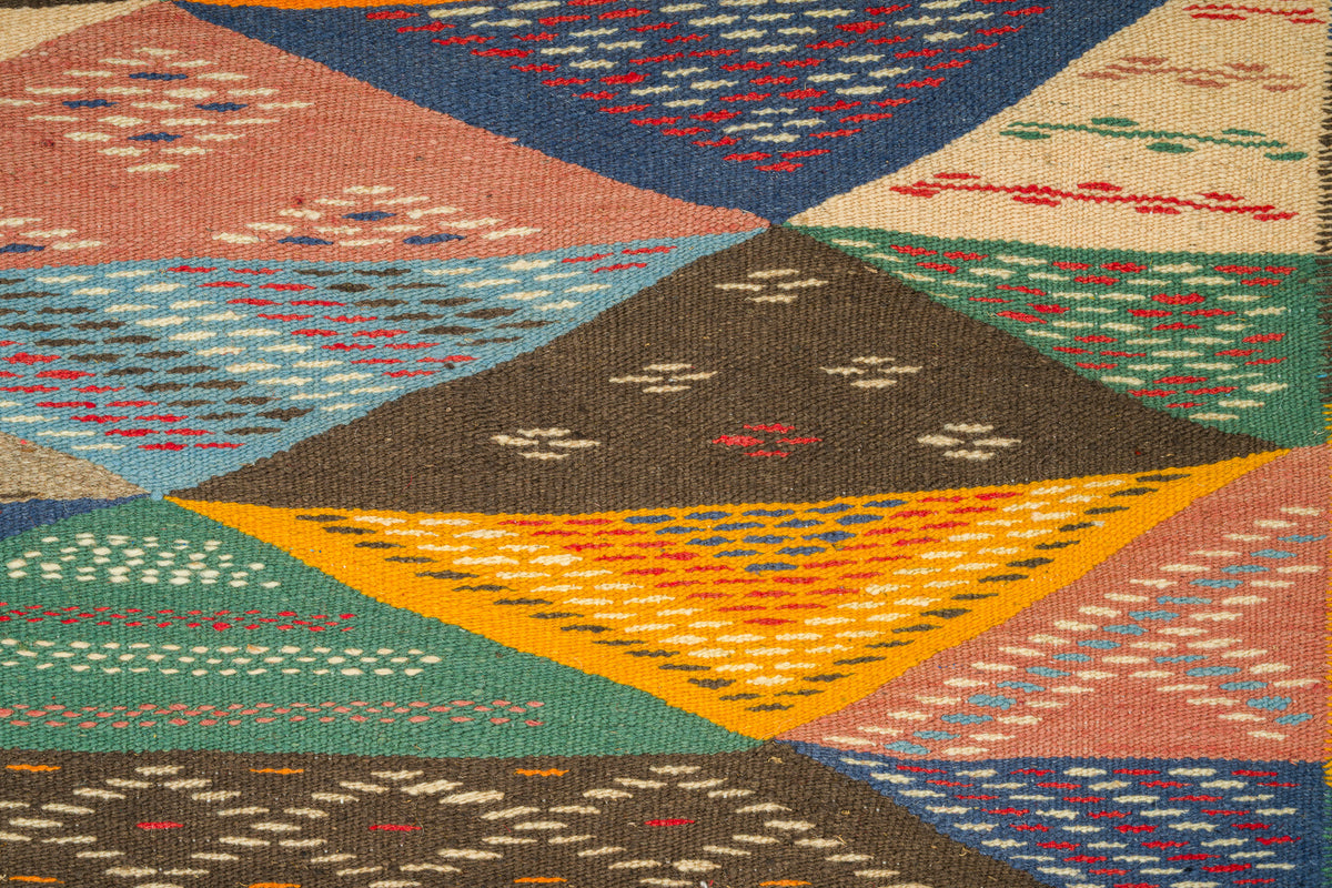 A close-up of a woven geometric moroccan rug with a patchwork design of triangular sections in blue, green, brown, orange, pink, and beige, each with embroidered diamond and zigzag patterns.