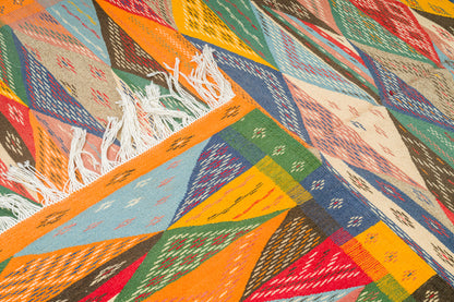 A close-up of a woven moroccan berber ru with a colorful geometric pattern in orange, blue, green, red, beige, and brown, featuring white fringe on one edge. 
