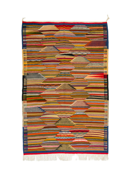 A rectangular woven moroccan rug or scarf with an intricate multicolor geometric pattern, featuring horizontal bands of red, blue, yellow, green, orange, and beige, finished with white fringe at the bottom.