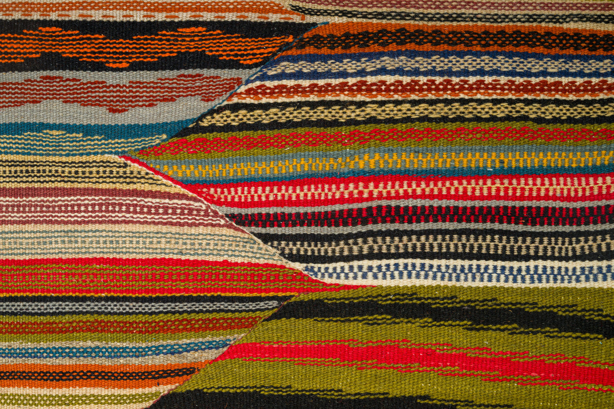 A close-up of a woven moroccan rug with horizontal and diagonal striped bands in orange, red, blue, beige, black, olive green, and brown.