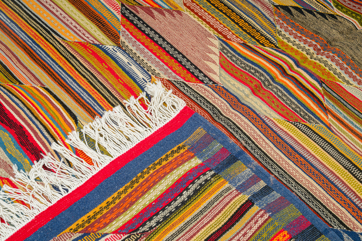 A close-up of a colorful woven moroccan rug with diagonal striped and geometric patterns in red, orange, yellow, blue, beige, and black, featuring white fringe on one end.
