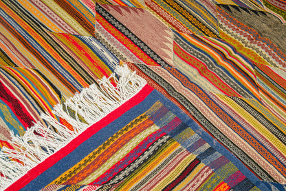 A close-up of a colorful woven moroccan rug with diagonal striped and geometric patterns in red, orange, yellow, blue, beige, and black, featuring white fringe on one end.