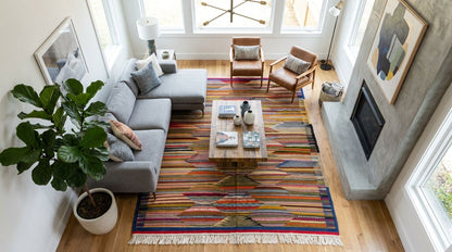 A high-angle view of a modern living room featuring a large, colorful geometric patterned moroccan area rug as the central decor piece, styled with a gray sectional sofa, wooden coffee table, and a potted fiddle-leaf fig plant.
