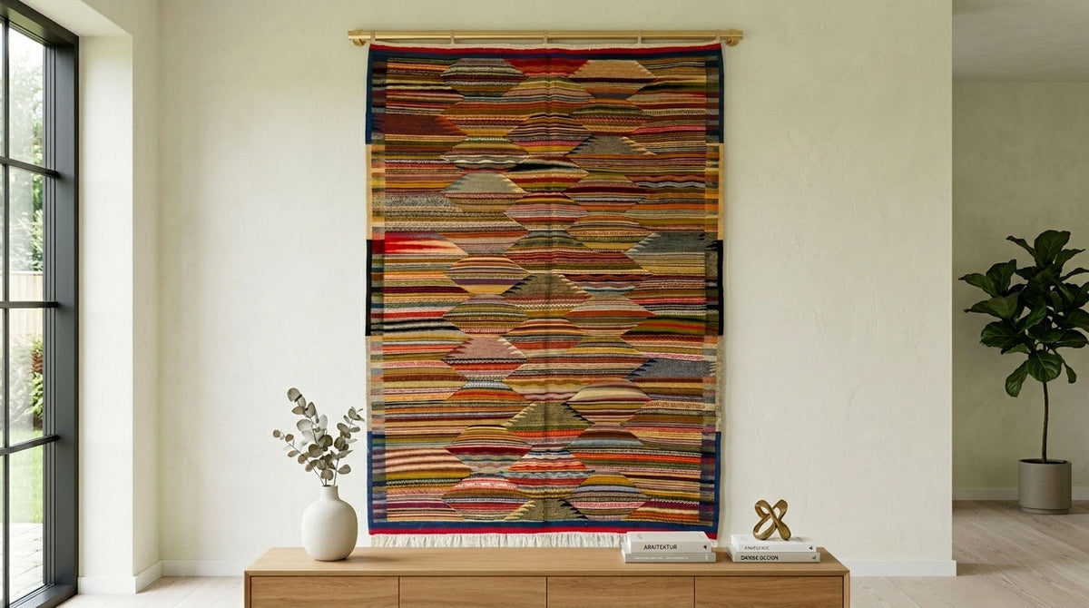 A large, multicolored moroccan rug used as  wall hanging or tapestry with horizontal layered stripes, suspended from a wooden rod in a modern, minimalist room with a wooden console table and a potted plant.