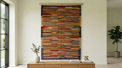 A large, multicolored moroccan rug used as  wall hanging or tapestry with horizontal layered stripes, suspended from a wooden rod in a modern, minimalist room with a wooden console table and a potted plant.