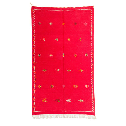 A red rectangular handwoven moroccan rug from akhnif with white fringe at the bottom, decorated with hand embroidered small multicolored geometric motifs and a white zigzag border.