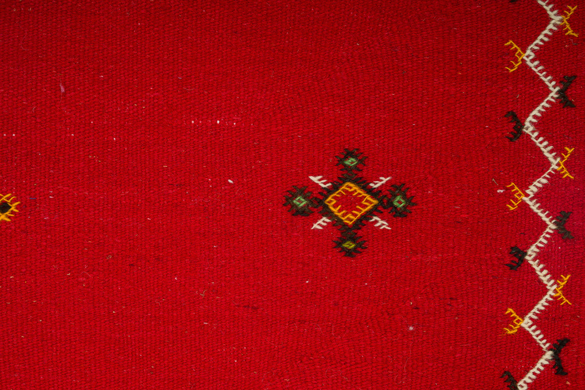 A close-up of a red handwoven moroccan rug with a small central hand embroidered geometric motif in black, green, yellow, and red, plus a vertical zigzag border in white, black, and yellow along the right edge.