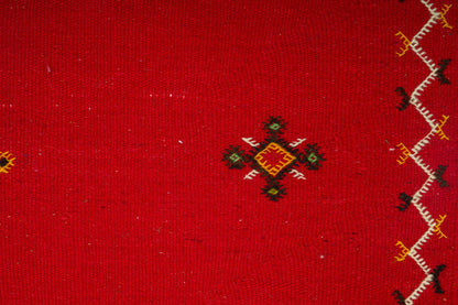 A close-up of a red handwoven moroccan rug with a small central hand embroidered geometric motif in black, green, yellow, and red, plus a vertical zigzag border in white, black, and yellow along the right edge.