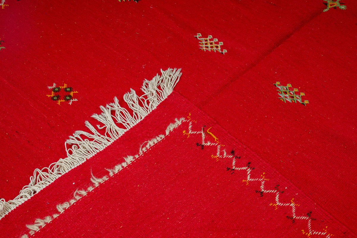 A close-up of a bright red textile or  handwoven moroccan rug showing white fringe along one edge and small multicolored hand embroidered geometric motifs scattered across the surface.
