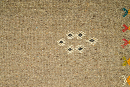 Close-up of a moroccan woven rug or textile from Akhnif in a warm beige/tan color, featuring a small hand embroidered central geometric diamond pattern in cream, black, and white, with partial multicolored motifs visible along the right edge.