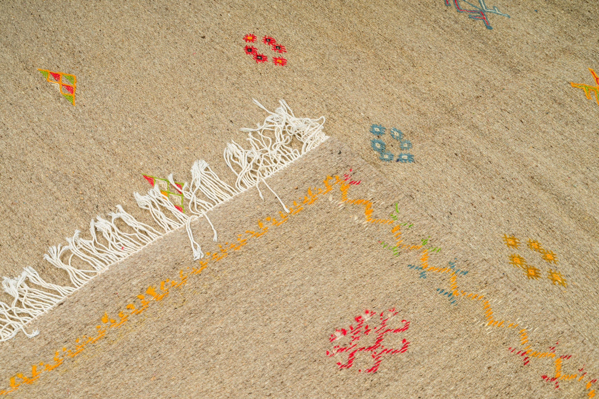 Close-up of a handwoven moroccan rug in a sandy beige color, featuring white fringe on one edge and small scattered embroidered floral motifs in red, yellow, and blue.