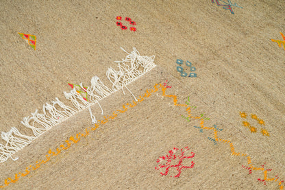 Close-up of a handwoven moroccan rug in a sandy beige color, featuring white fringe on one edge and small scattered embroidered floral motifs in red, yellow, and blue.