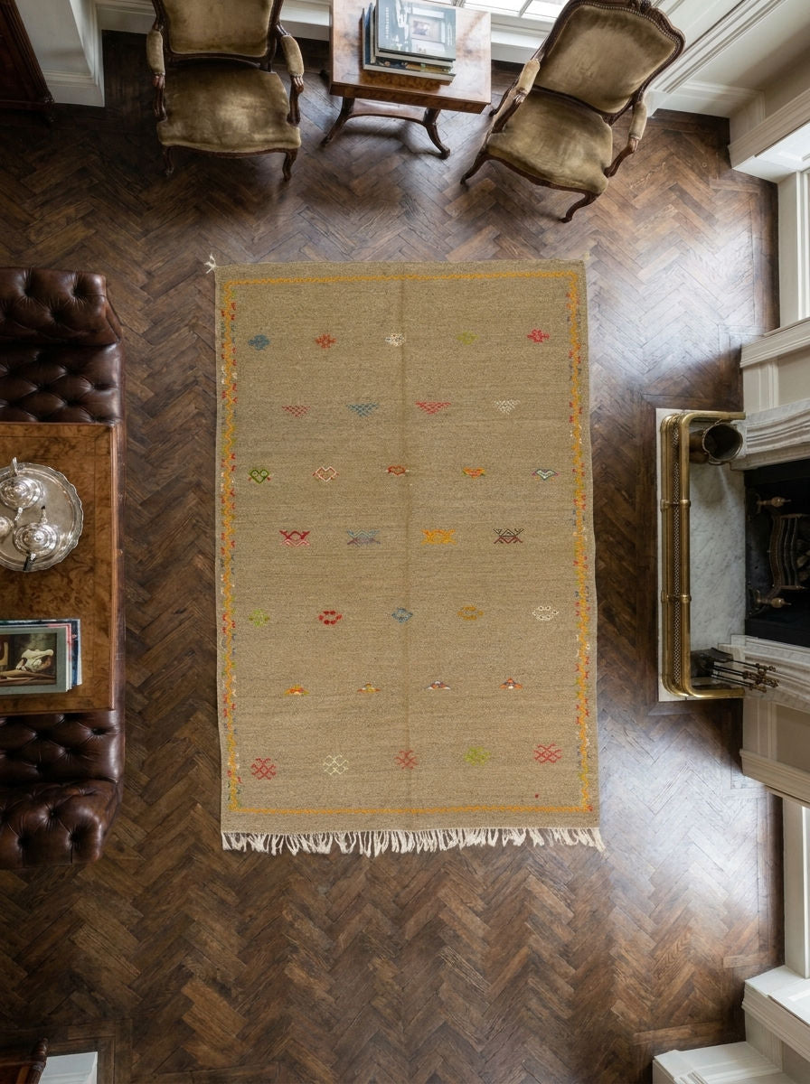 A rectangular beige moroccan area rug with a handwoven texture, colorful hand embroidered geometric motifs, and fringe along the bottom edge, shown from a top-down perspective in a furnished room with dark wood herringbone flooring.