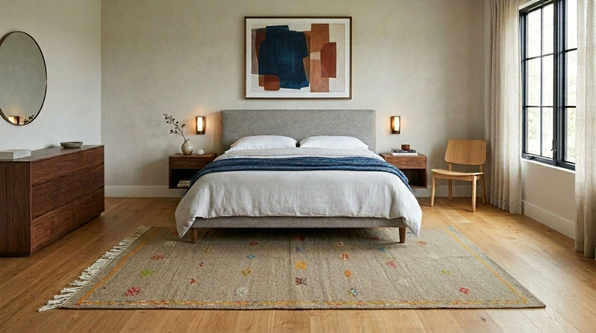 A styled bedroom interior featuring a bed with white bedding and a blue throw, set on a patterned moroccan area rug over light wood flooring. The room includes a gray upholstered headboard, matching nightstands, a wooden chair, a dresser, and an abstract painting on the wall.