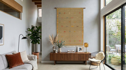 A large handwoven moroccan wall hanging  rug with a natural beige base and subtle multicolor hand embroidered motifs, displayed in a bright, modern living room above a wooden console table.