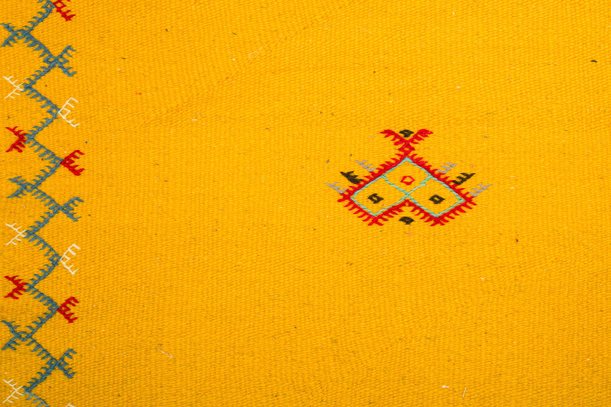 Yellow  moroccan rug handwoven and hand embroidered with a small red, black, and gray geometric motif near the center and a blue and red embroidered border along the left edge.
