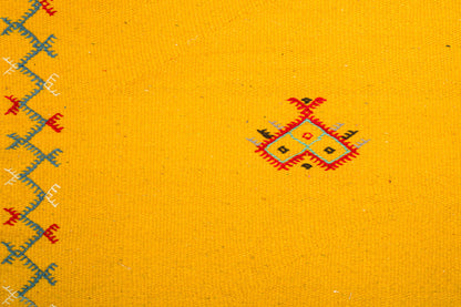 Yellow  moroccan rug handwoven and hand embroidered with a small red, black, and gray geometric motif near the center and a blue and red embroidered border along the left edge.
