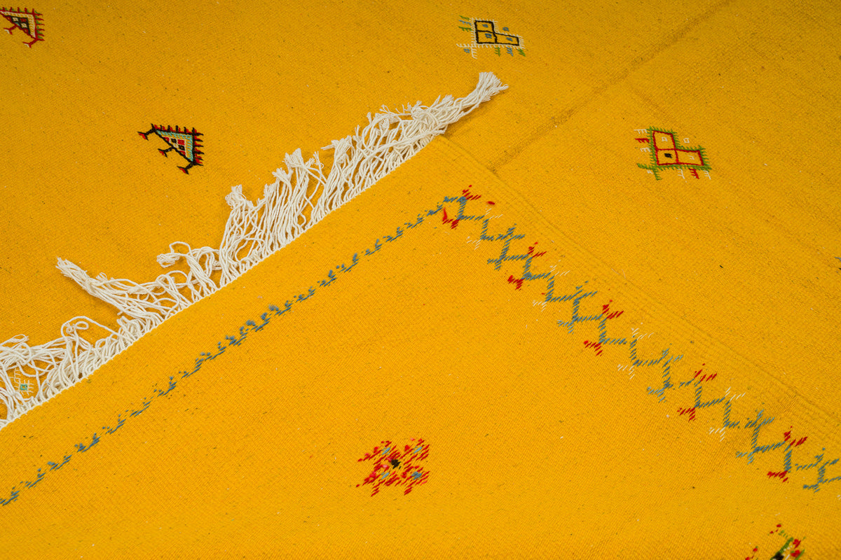 A close-up of a bright yellow moroccan rug or textile with white fringe, embroidered geometric motifs, and a stitched zigzag border.