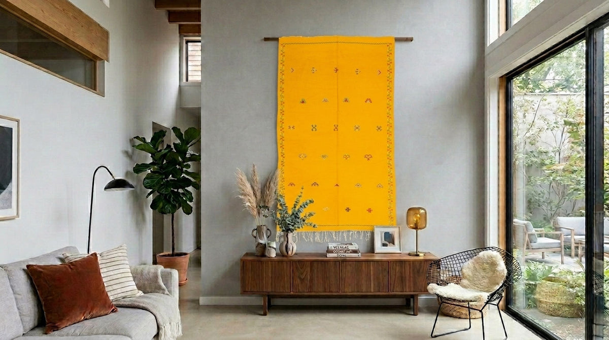 A bright yellow moroccan woven wall hanging with small embroidered motifs is displayed in a modern living room setting above a wooden console table.