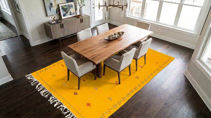 A bright yellow moroccan area rug with fringe and small hand embroidered floral motifs, styled under a wooden dining table and six gray upholstered chairs in a modern dining room with dark wood floors and large windows.