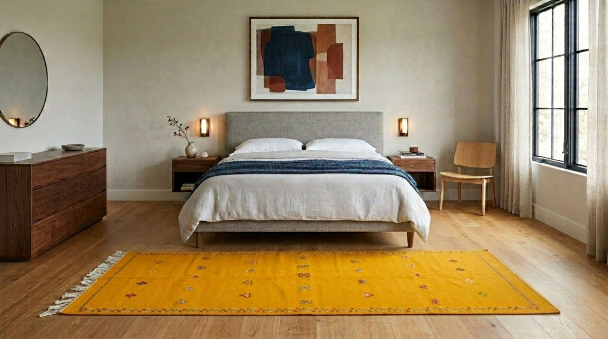 A bright yellow moroccan area rug laid out on a light wood floor in a modern, neutral-toned bedroom with a bed, wooden nightstands, and a window.