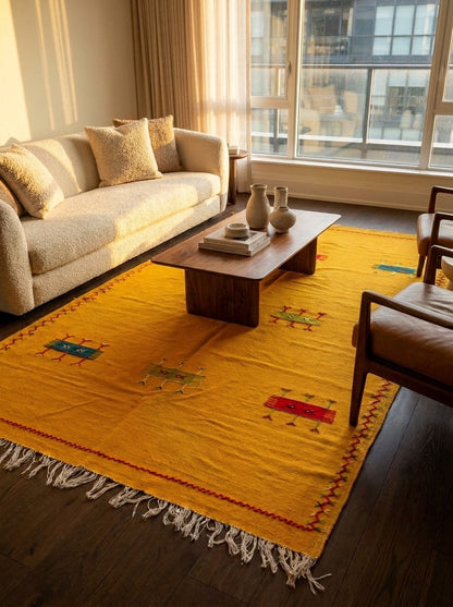 A bright yellow moroccan area rug with white fringe and small hand embroidered colorful geometric motifs, styled in a modern living room with a beige sofa, wooden coffee table, and large windows.
