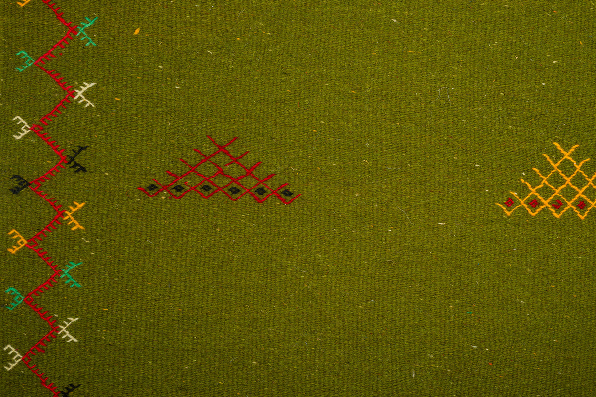 Close-up of olive green moroccan rug with embroidered geometric patterns in red, yellow, black, and teal along the left edge and center.