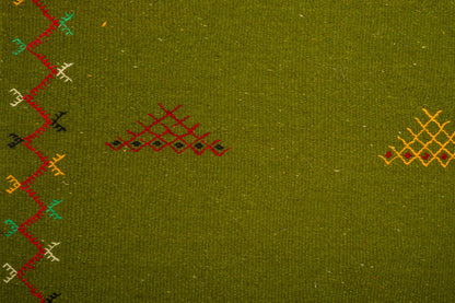 Close-up of olive green moroccan rug with embroidered geometric patterns in red, yellow, black, and teal along the left edge and center.